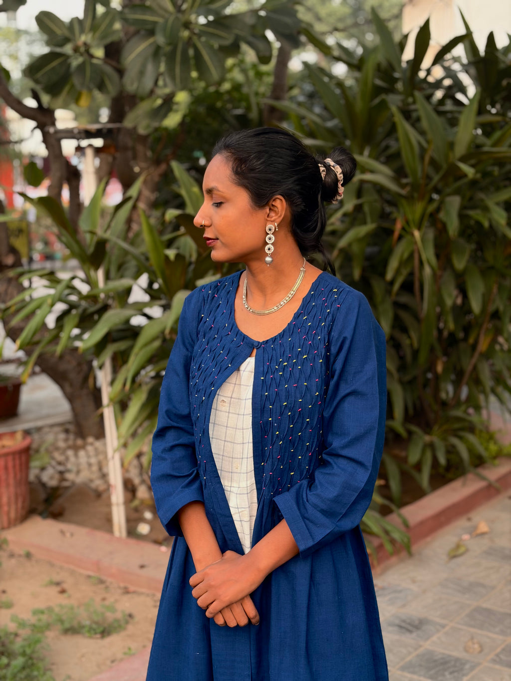 Woman in a blue shrug and white outfit standing outdoors with greenery in the background