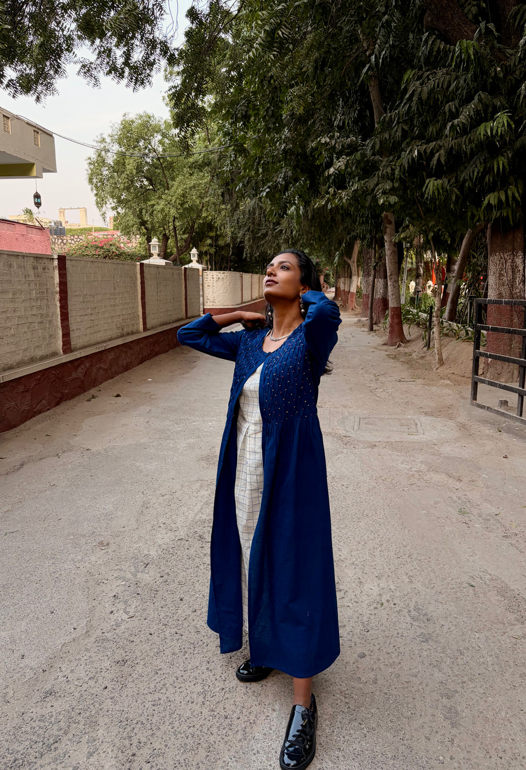 Woman in a blue shrug and white dress standing on a street with trees and buildings in the background