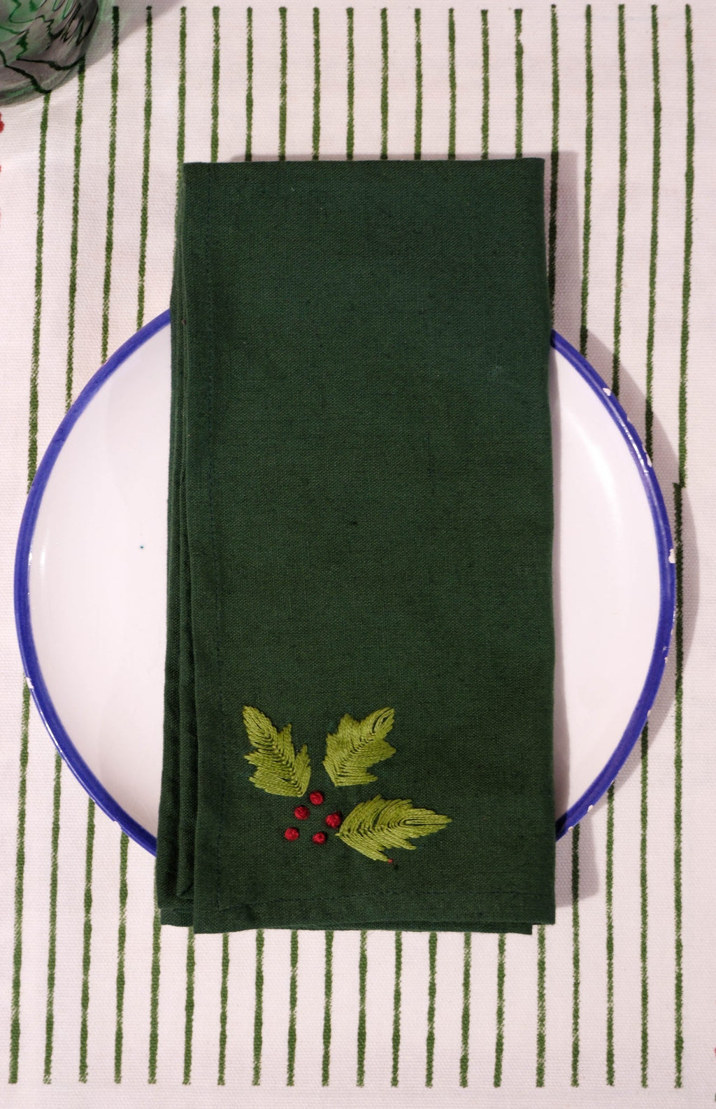 Green hand embroidered napkin with holly leaves on a white plate against a striped placemat