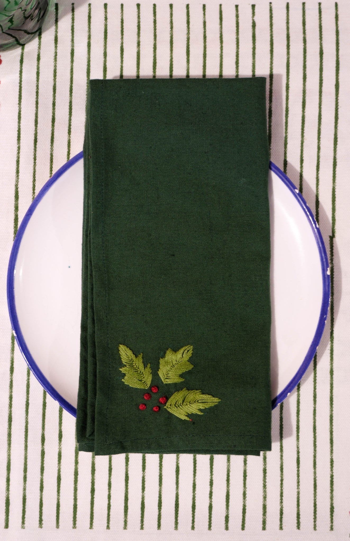 Green hand embroidered napkin with holly leaves on a white plate against a striped placemat