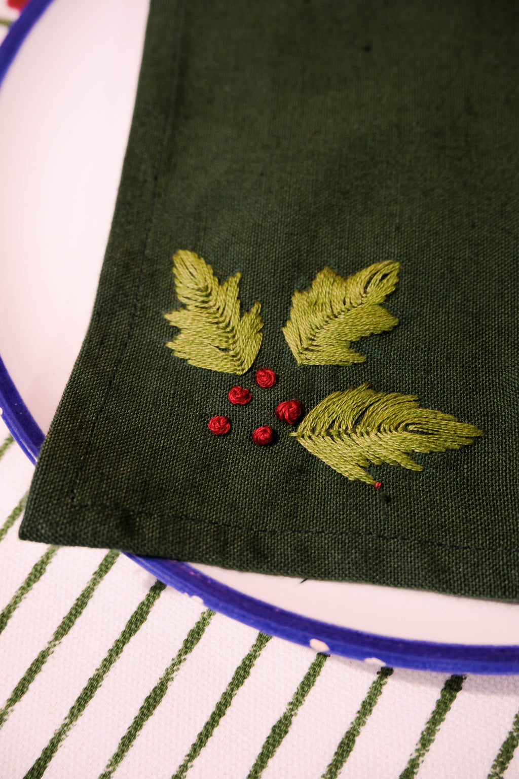 Green hand embroidered napkin with holly leaves on a white plate against a striped placemat